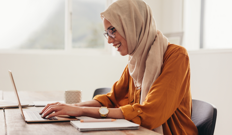 Woman wearing hijab working on laptop at home. Woman working from home using laptop and smiling.